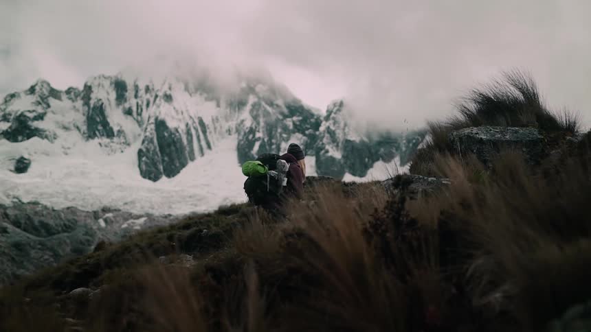 A hiker with a large backpack pauses on a windy, grassy ridge with a massive snow-capped mountain in the background