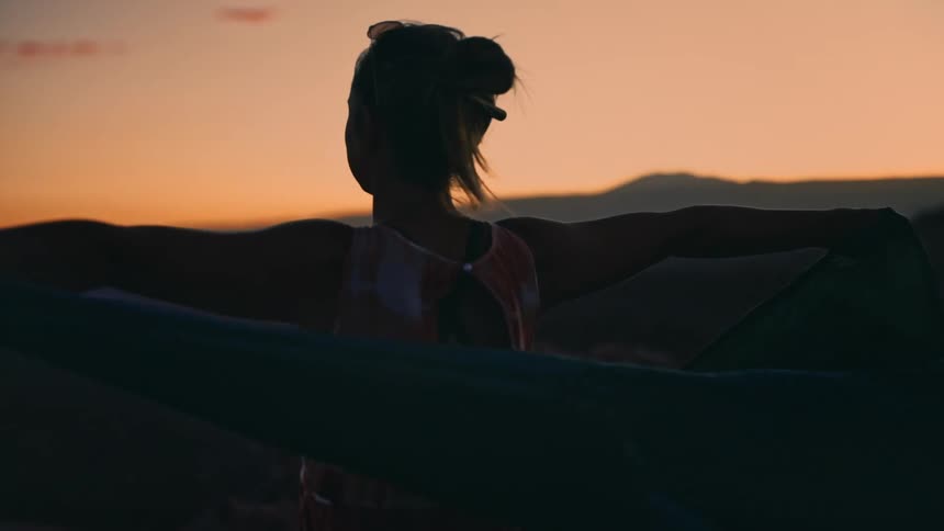 Slow motion shot of a woman's silhouette holding a large flowing fabric in the wind during a beautiful desert sunset