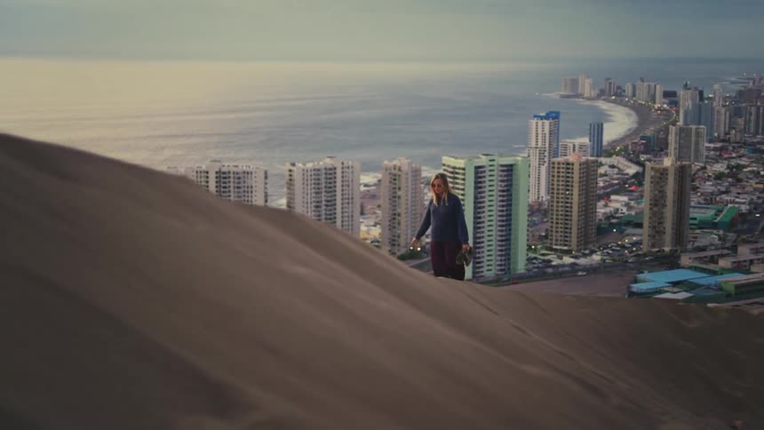 A woman walks in slow motion up a giant sand dune with a breathtaking view of a modern coastal city at dusk