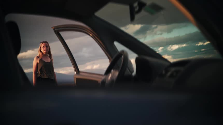 A young woman stands outside her car at golden hour, contemplating the cloudy sky and scenic landscape, as seen from inside the vehicle
