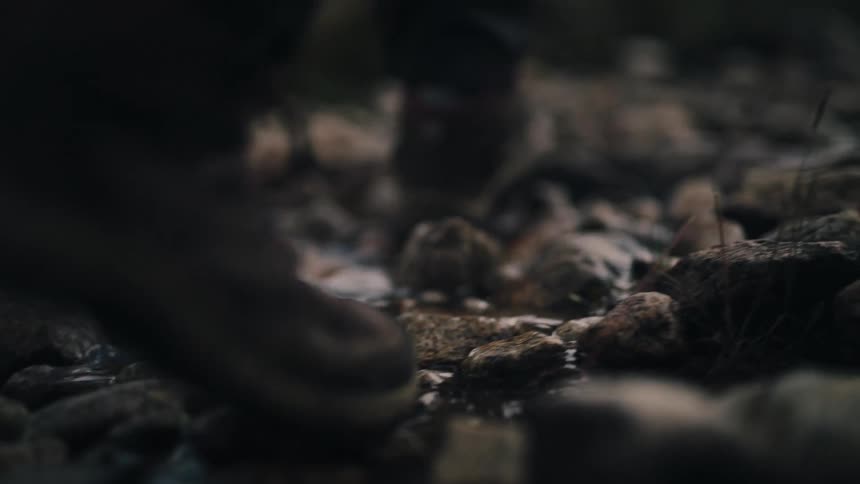 Low-angle slow-motion shot of a hiker's boots moving through a rocky, shallow stream