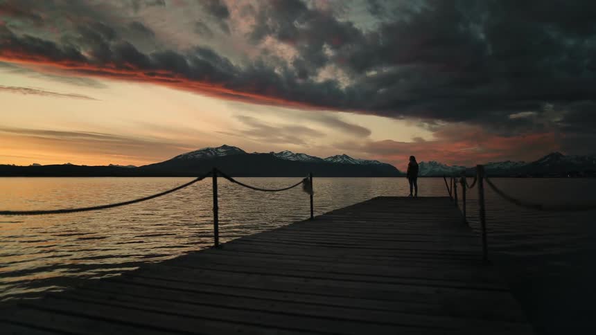 A person's silhouette stands at the end of a wooden pier, contemplating a dramatic sunset over a tranquil lake and distant mountains