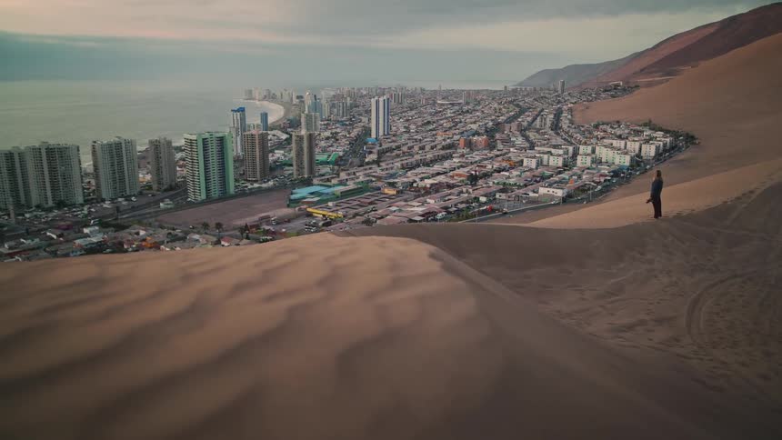 A lone man walks up a vast sand dune to overlook a sprawling coastal city nestled between the desert and the ocean