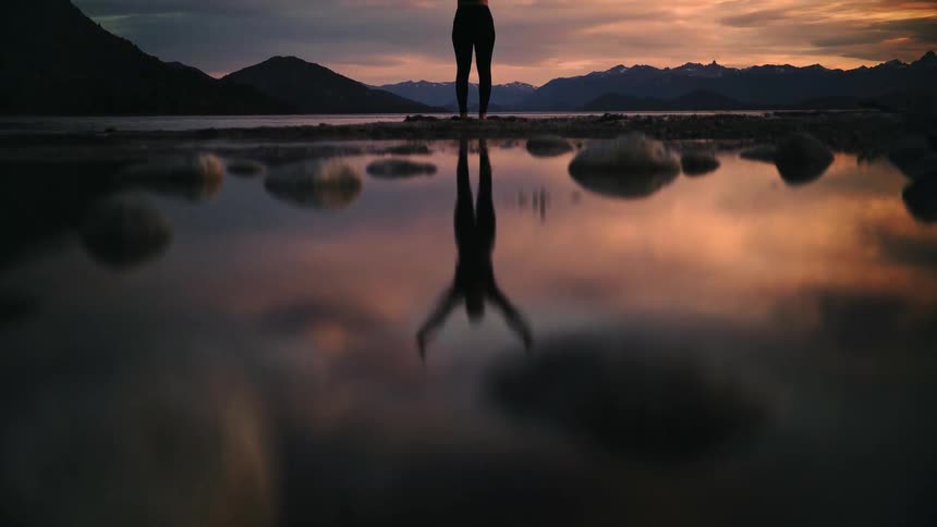 A lone figure stands on a rocky shore, their silhouette perfectly reflected in the calm lake water against a dramatic sunset sky