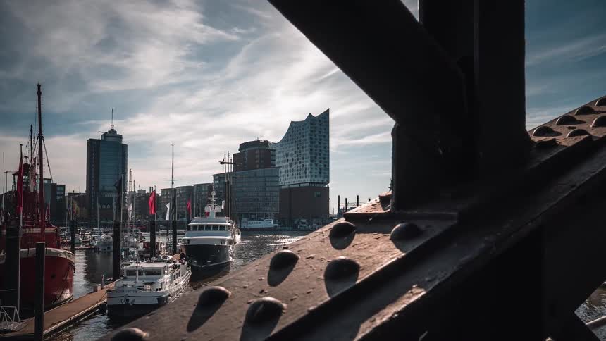 Dynamic time-lapse from behind a steel bridge structure reveals the iconic Elbphilharmonie and bustling boat traffic in Hamburg's harbor on a sunny day