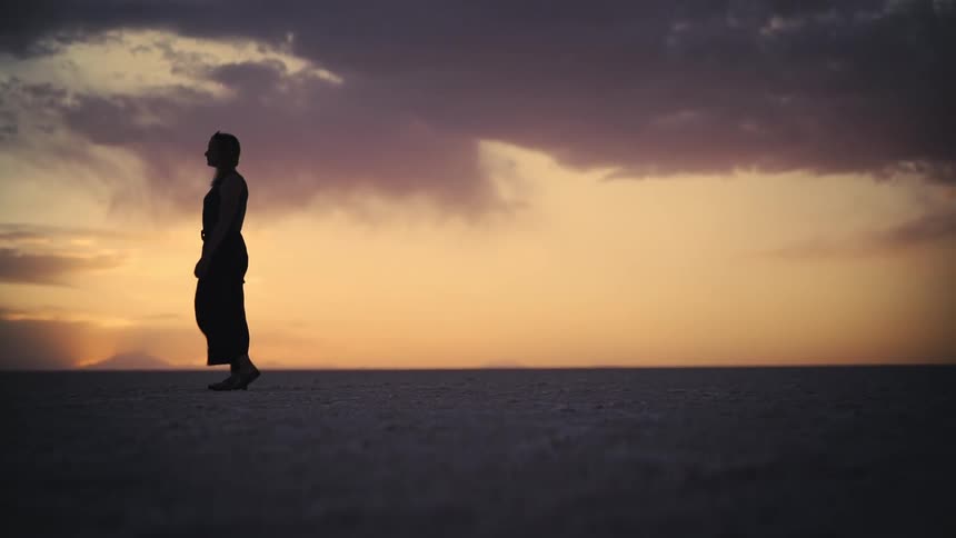 A cinematic slow motion shot captures the silhouette of a person walking across a vast, barren landscape at sunset