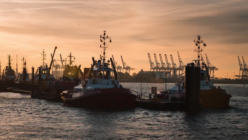 Silhouetted tugboats and industrial vessels float peacefully in a busy port during a golden sunset
