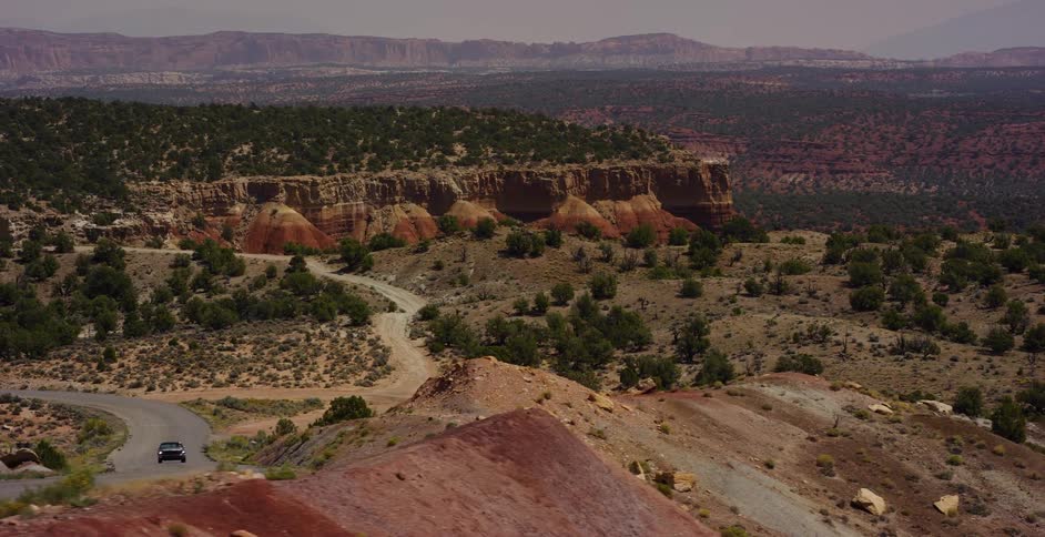 A slow aerial pullback reveals a black SUV driving along a winding desert road through a vast and colorful canyon landscape