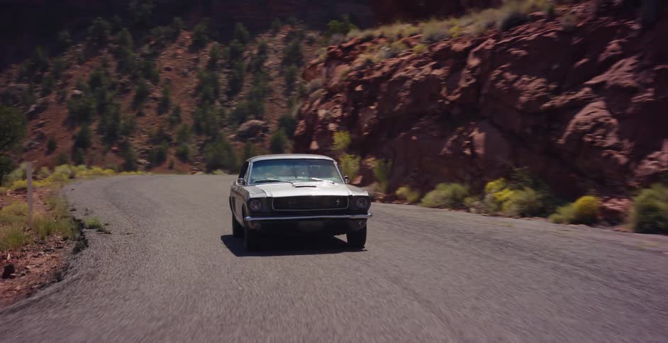 A classic silver Ford Mustang drives along a winding road through a red rock canyon on a sunny day