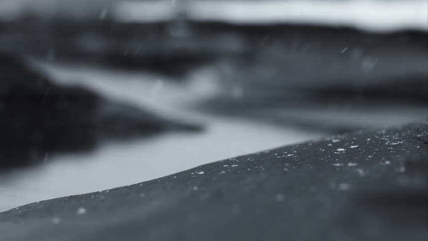 A tranquil, black and white slow-motion shot shows snow falling gently on a dark, rocky shore next to calm water