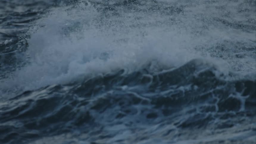 Turbulent dark blue ocean water churns as waves crest and crash in a powerful slow-motion display