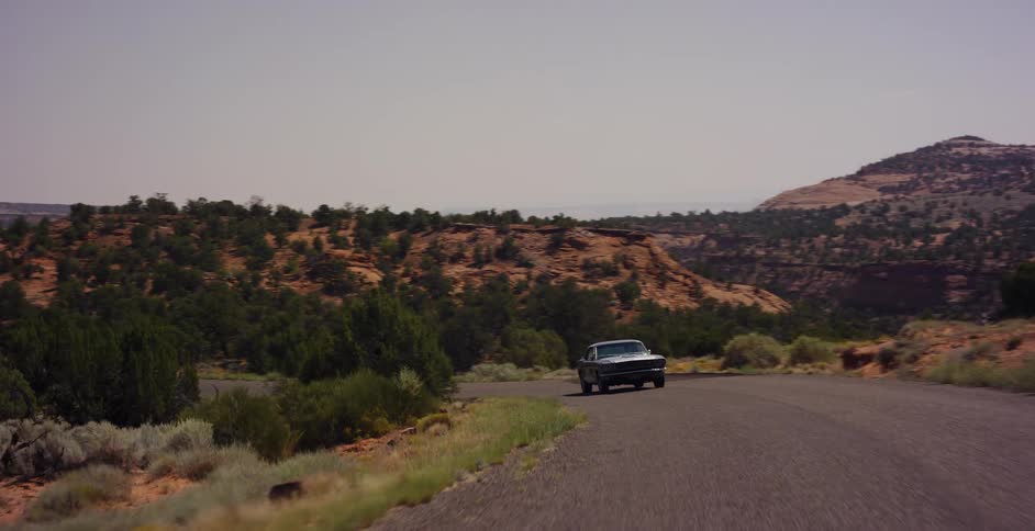 A classic muscle car cruises down a scenic, winding road through the vast desert canyonlands