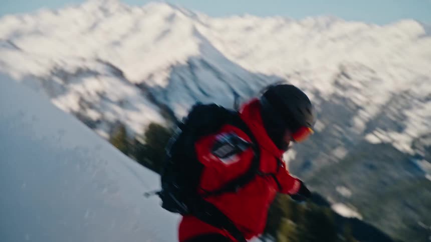 A snowboarder in a red jacket carves down a steep, snowy mountain in slow motion, revealing a picturesque valley below