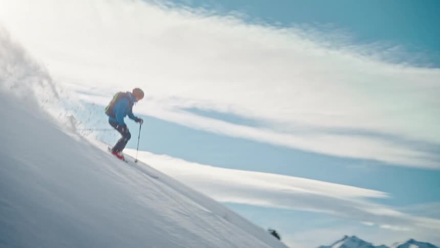 An extreme skier descends a steep, untouched powder snow slope on a beautiful sunny day in the mountains in slow motion