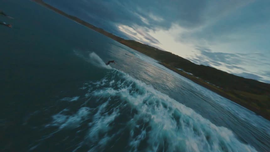 An exciting FPV drone chases a surfer riding a rolling wave along the coastline during a dramatic sunset
