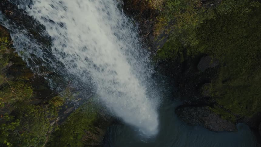 An aerial top-down view shows a powerful waterfall cascading down a lush green cliff into a tranquil pool below