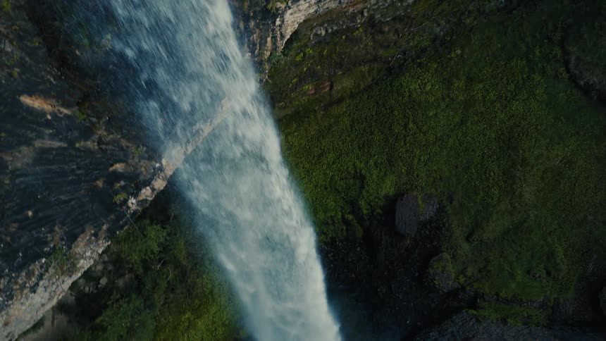 An amazing top-down aerial shot reveals a powerful waterfall crashing down from a cliff into a pool surrounded by lush greenery