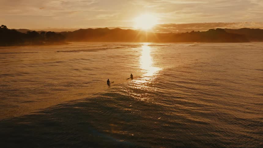 Two surfers peacefully float on their boards in the ocean, silhouetted against a magnificent golden sunset over distant mountains