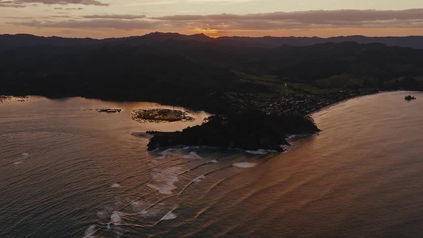 An aerial view reveals a stunning coastal peninsula and town as gentle waves roll in during a golden sunset