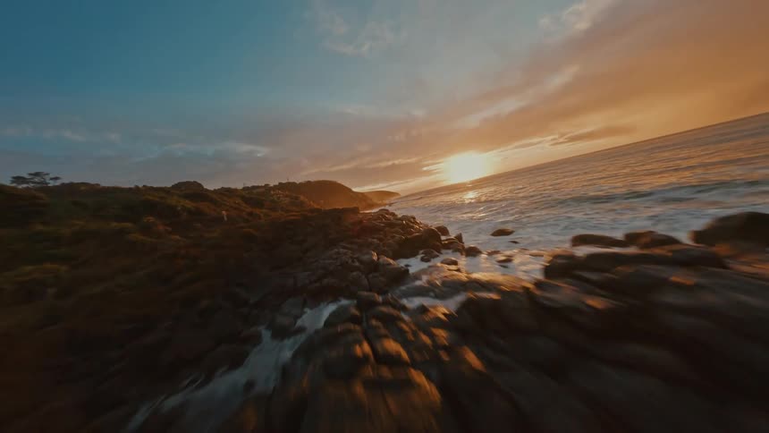 An FPV drone flies low over a rocky coastline as waves crash against the shore during a golden sunset