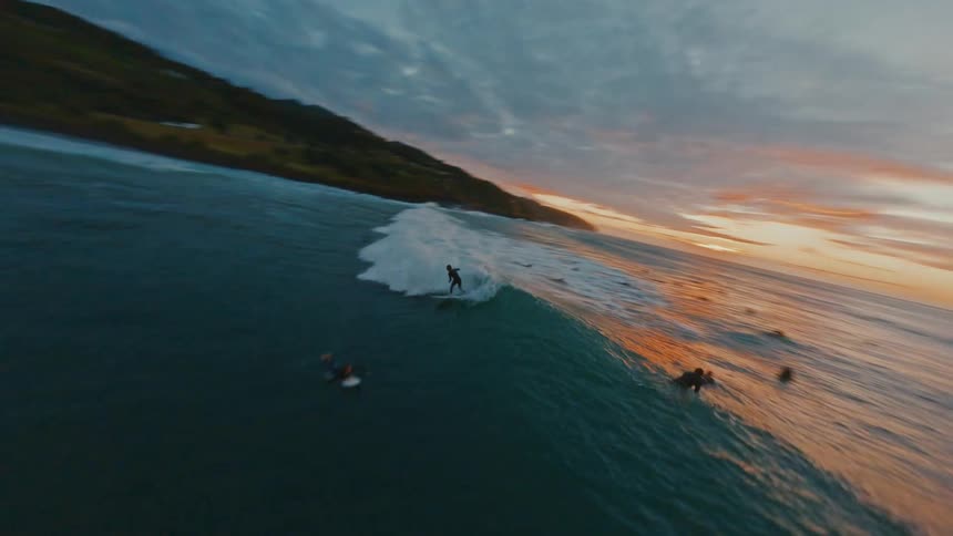 An FPV drone follows a surfer catching and riding a wave along the coast during a beautiful, moody sunset