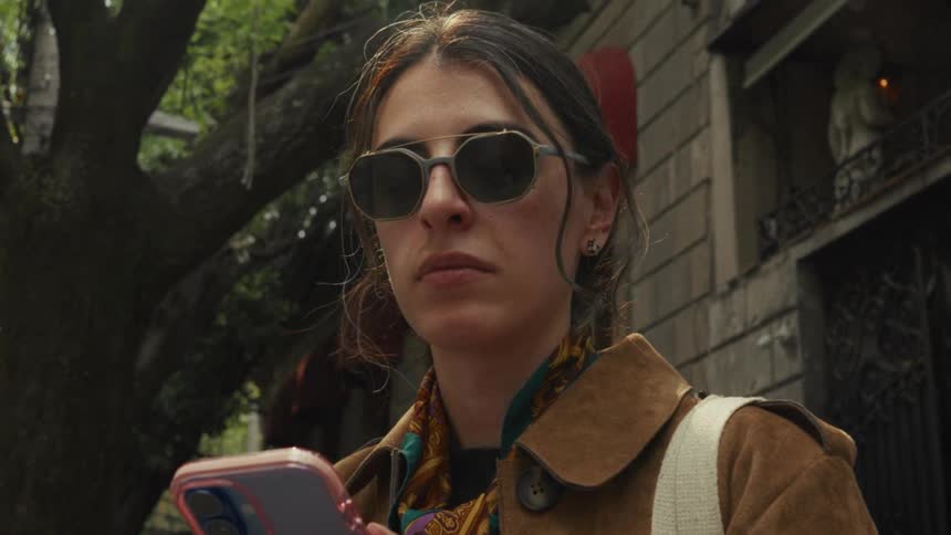 A woman in a brown coat and sunglasses pauses on a tree-lined city street, looking up from her phone with a thoughtful expression
