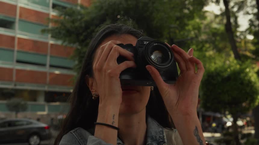A female photographer holds a camera to her face and adjusts the focus while standing on a city street