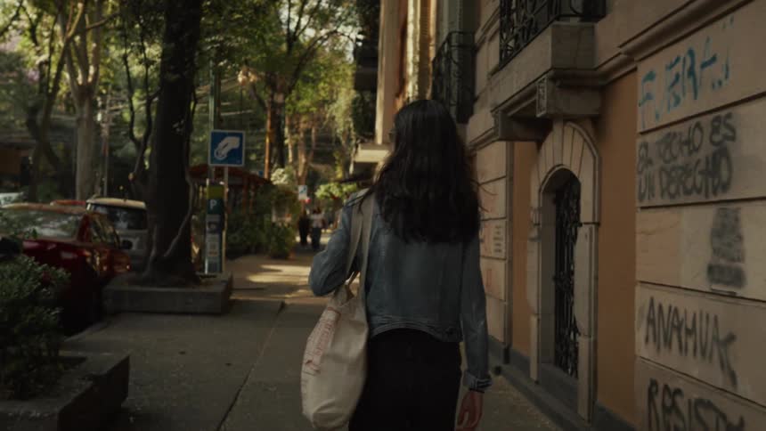 A young woman with long dark hair walks down a leafy city sidewalk in slow motion