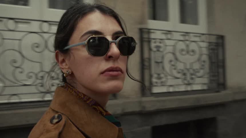 A stylish woman in sunglasses and a brown jacket walks down a city street in slow motion, stopping to look around