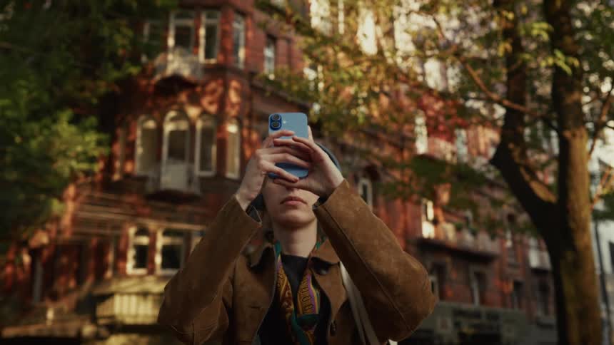 A young woman wearing headphones takes a photo of a beautiful old building with her smartphone while standing on a city street at sunset