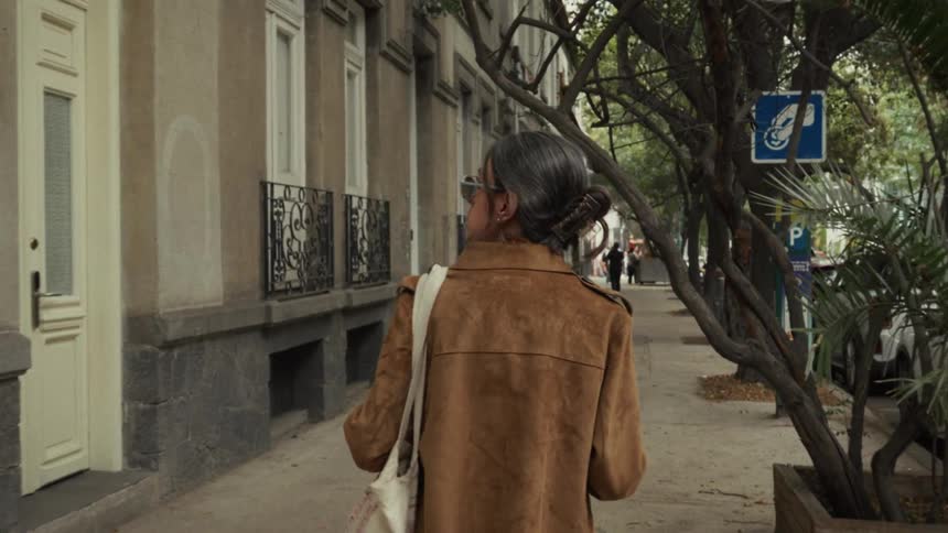 Cinematic slow-motion shot from behind of a woman in a brown suede jacket walking down a tree-lined city sidewalk