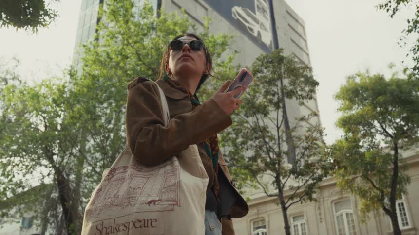 A woman wearing sunglasses walks through a city, stopping to check her phone while looking up at a modern building