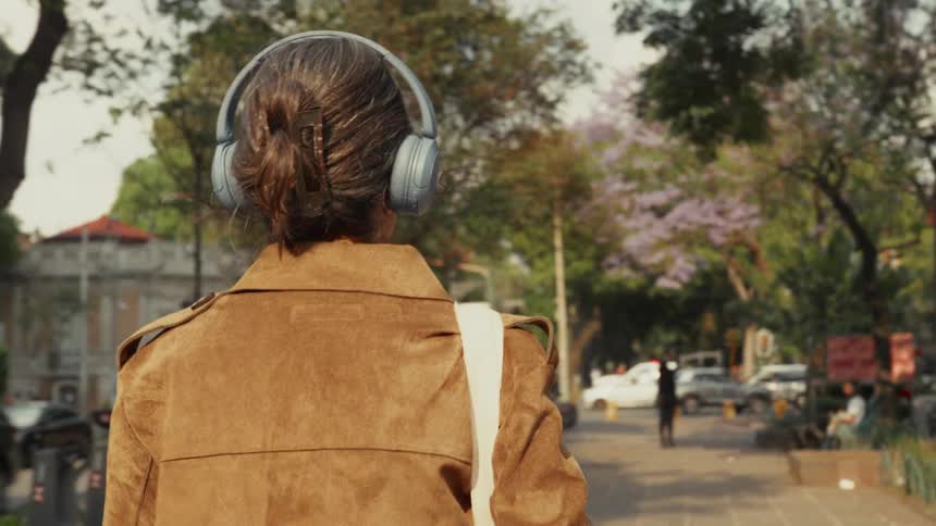 A stylish woman wearing headphones and sunglasses walks down a tree-lined city street on a sunny day