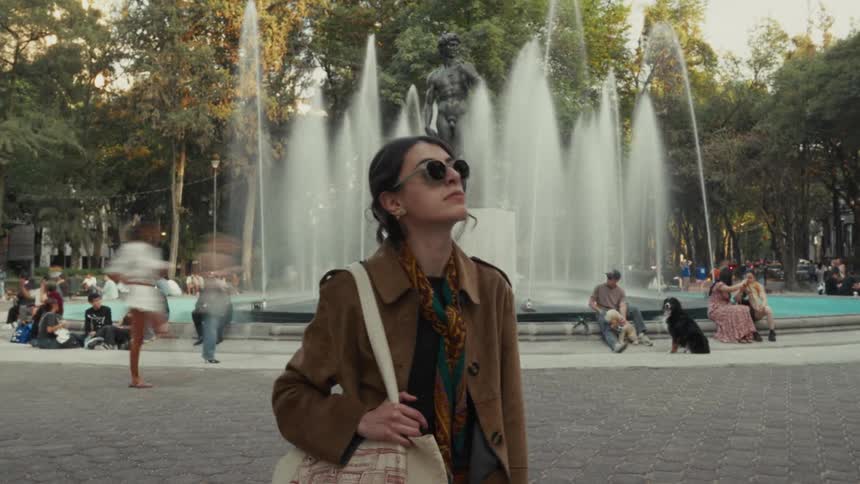 Time-lapse of a stylish woman standing still in a bustling city park in front of a fountain