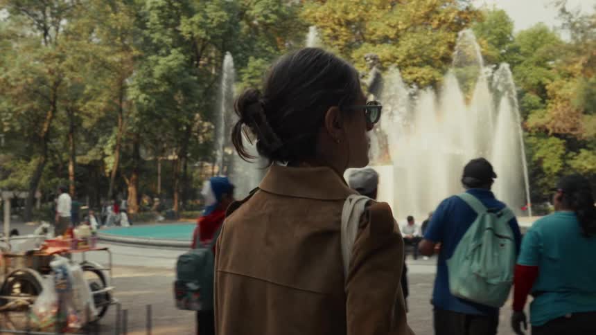A woman in a suede jacket and sunglasses walks through a bustling city park past a large fountain