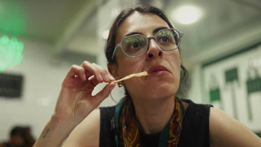 A contemplative woman with glasses eats a tortilla chip in slow motion while sitting in a diner