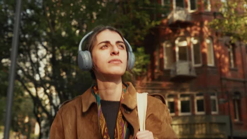 A thoughtful young woman wearing headphones enjoys a peaceful stroll on a sunlit city street in slow motion