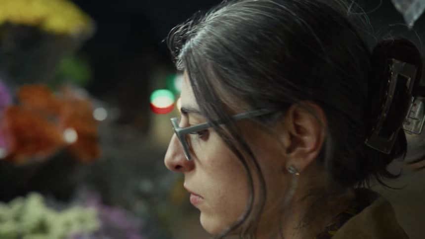 A woman with glasses thoughtfully admires a colorful flower stand on a city street at night