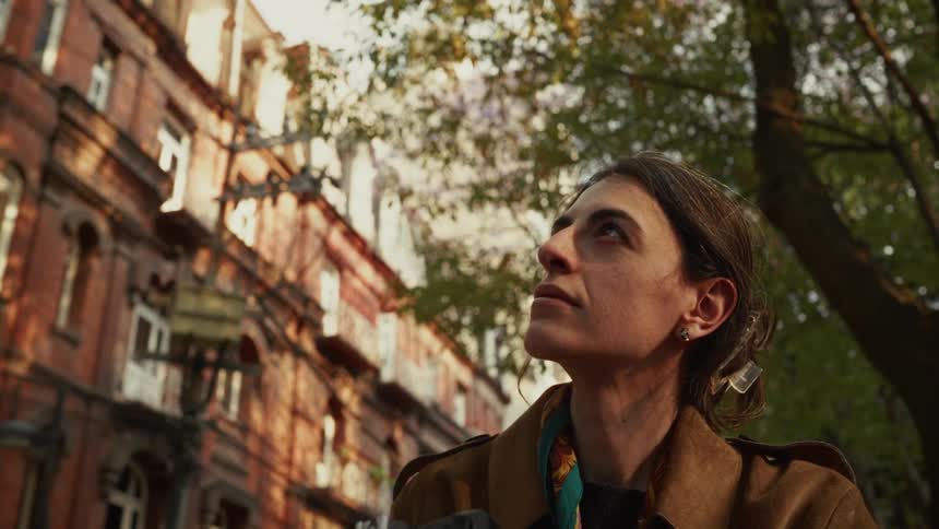 A thoughtful female photographer takes pictures of old city architecture on a tree-lined street during golden hour