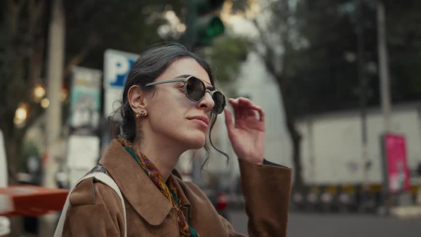 A stylish woman in sunglasses waits patiently on a bustling city street corner in slow motion