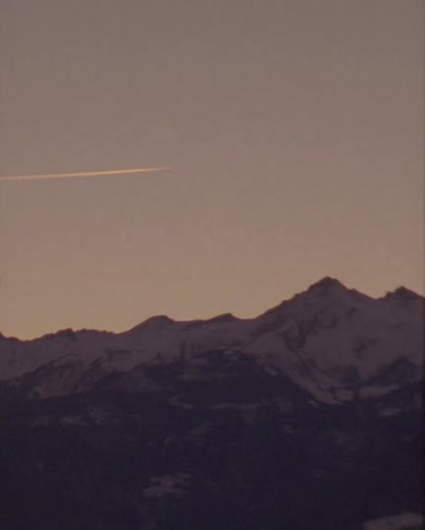 A slow pan shot shows a jet contrail streaking across the sky above a snowy mountain range at dusk