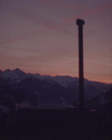 The silhouette of a chimney rises against a purple and pink twilight sky over a snowy mountain range
