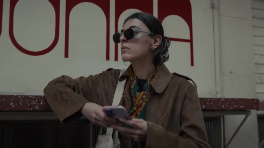 A chic woman wearing sunglasses and a brown coat leans against a building, looks at her phone, and then waits patiently on a city street