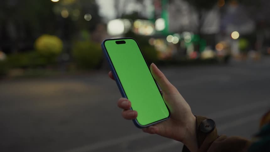 Over-the-shoulder shot of a person holding a smartphone with a chroma key green screen on a city street at night