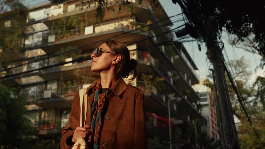A fashionable young woman in sunglasses pauses to enjoy the golden hour sunlight on a city street before walking away