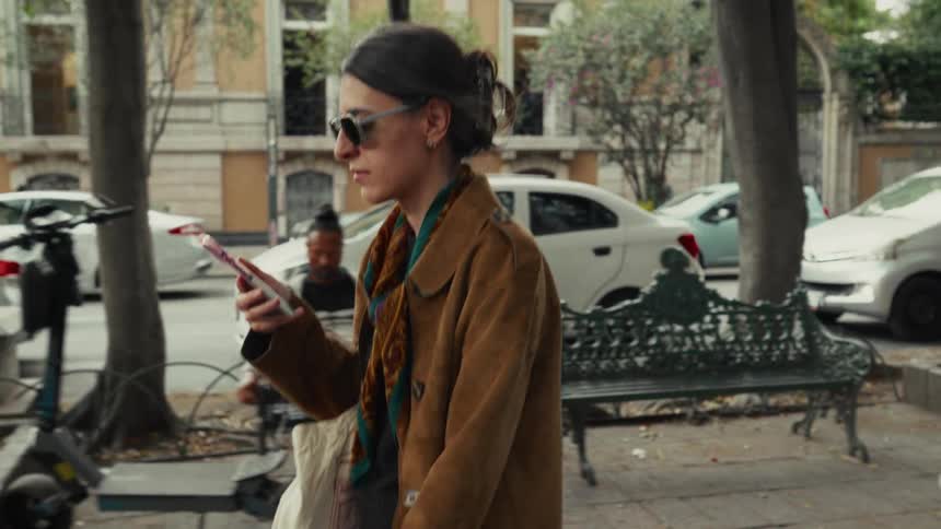 A stylish woman in sunglasses walks down a busy city street in slow motion while holding her smartphone