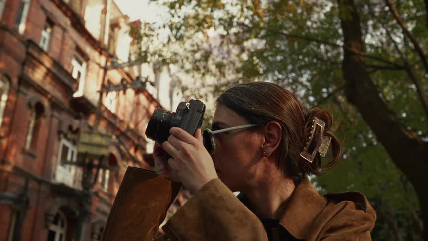 Low-angle slow-motion shot of a stylish woman in sunglasses taking photos of city architecture in the warm afternoon light