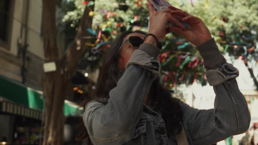 A young woman wearing sunglasses and a denim jacket films colorful festival ribbons on a tree with her smartphone in slow motion