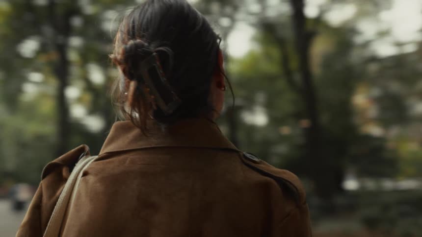 A contemplative young woman in a stylish brown jacket walks through a bustling city park, admiring the scenery and a large water fountain in slow motion