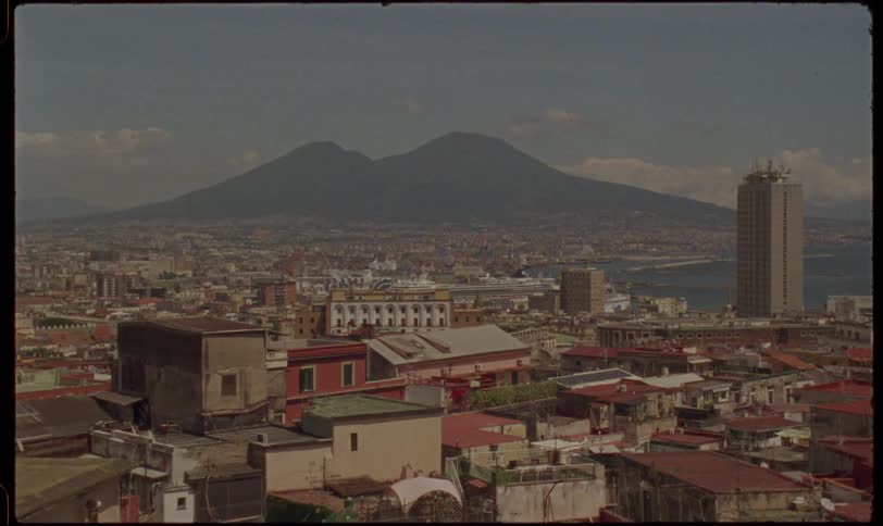 A vintage film look captures a slow zoom over the historic rooftops of Naples, Italy, towards the iconic Mount Vesuvius on a sunny day
